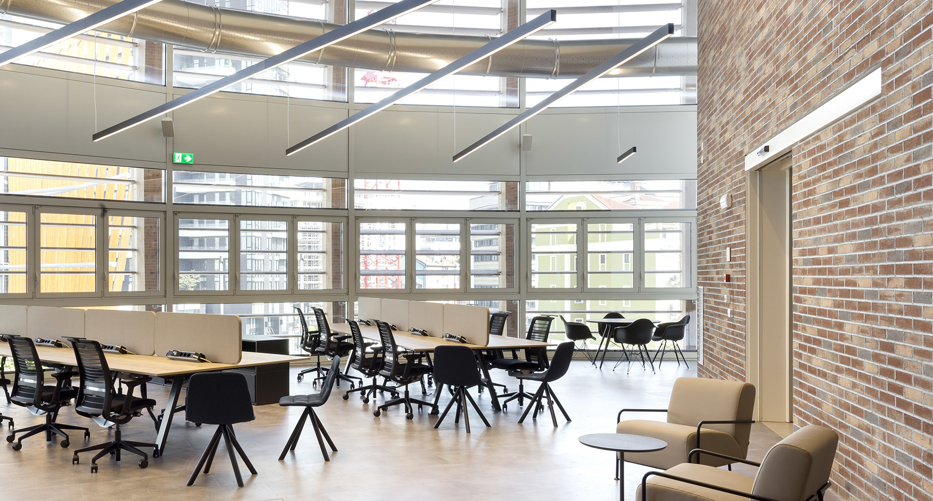 Modern open-plan office with Artemide linear pendant lights on ceiling, workstations with black chairs, exposed brick wall, large windows
