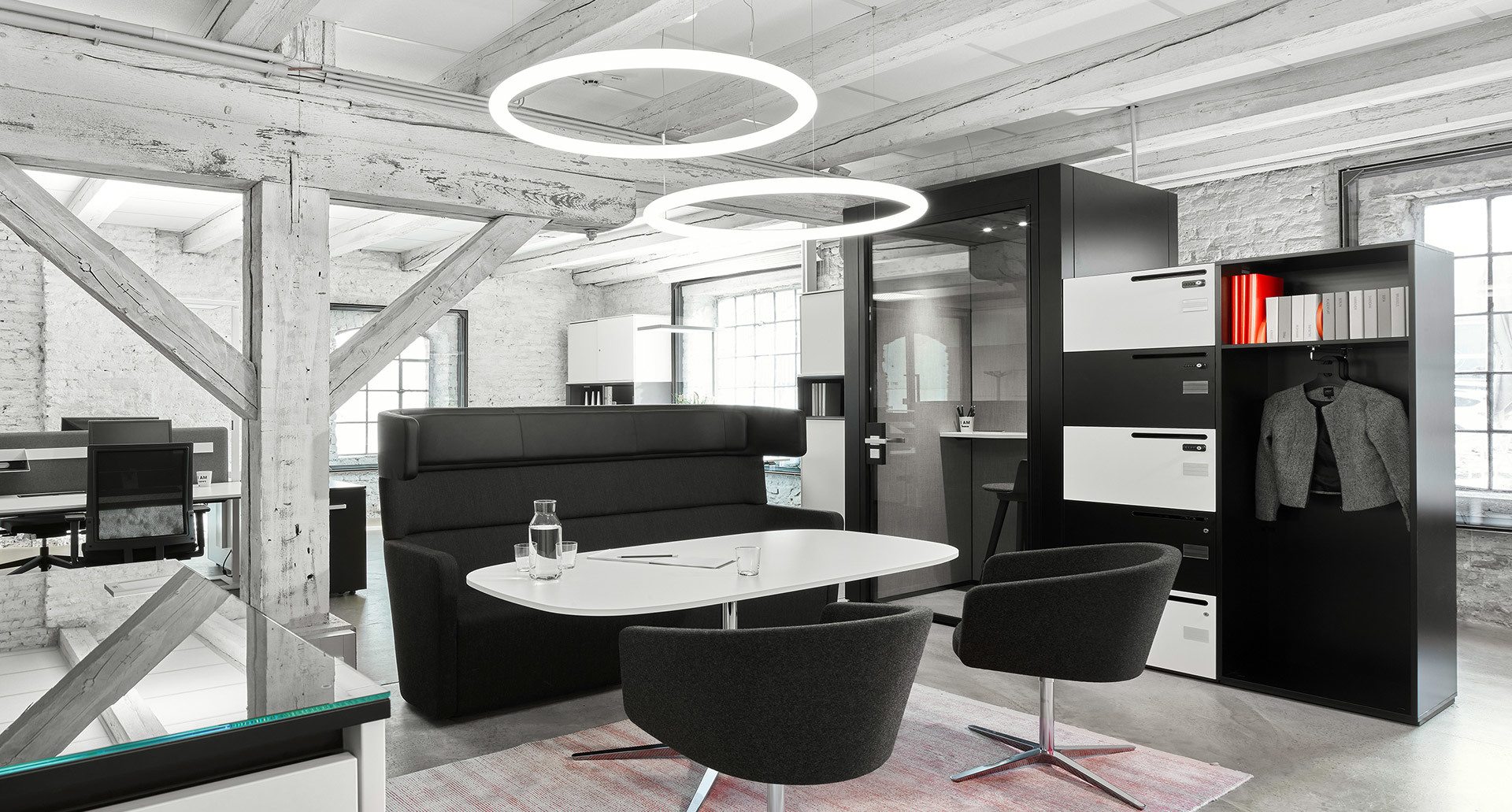 Modern office lounge with two Artemide circular ring pendant lights illuminating black seating, oval table, and industrial brick walls.