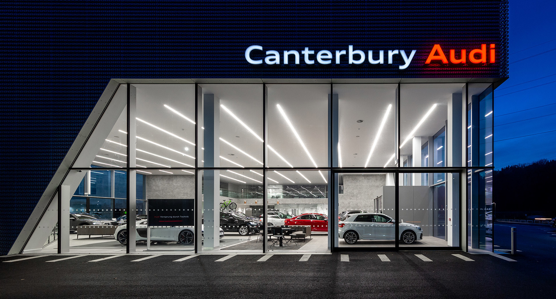Canterbury Audi showroom at night with linear LED ceiling lights illuminating luxury vehicles behind floor-to-ceiling glass facade