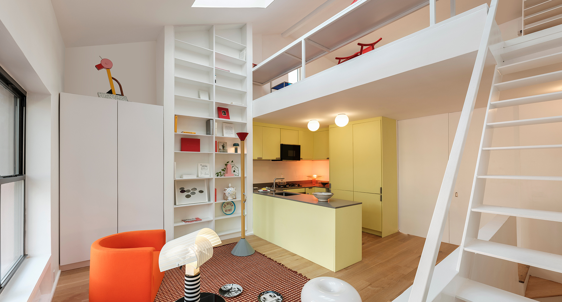 Modern loft interior with Artemide Snoopy and Tolomeo lamps, yellow kitchen, mezzanine level, white shelving, and orange armchair.