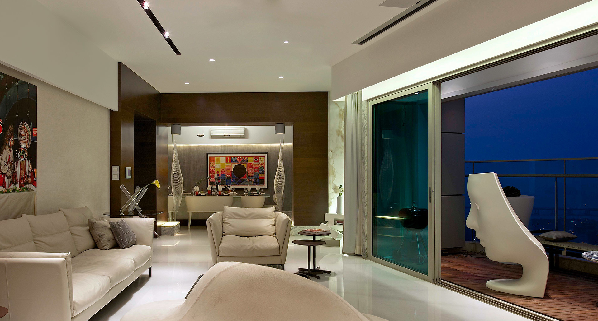 Modern living room with recessed ceiling lights and track lighting, cream sofas, open glass doors to night balcony, and a sculptural white chair
