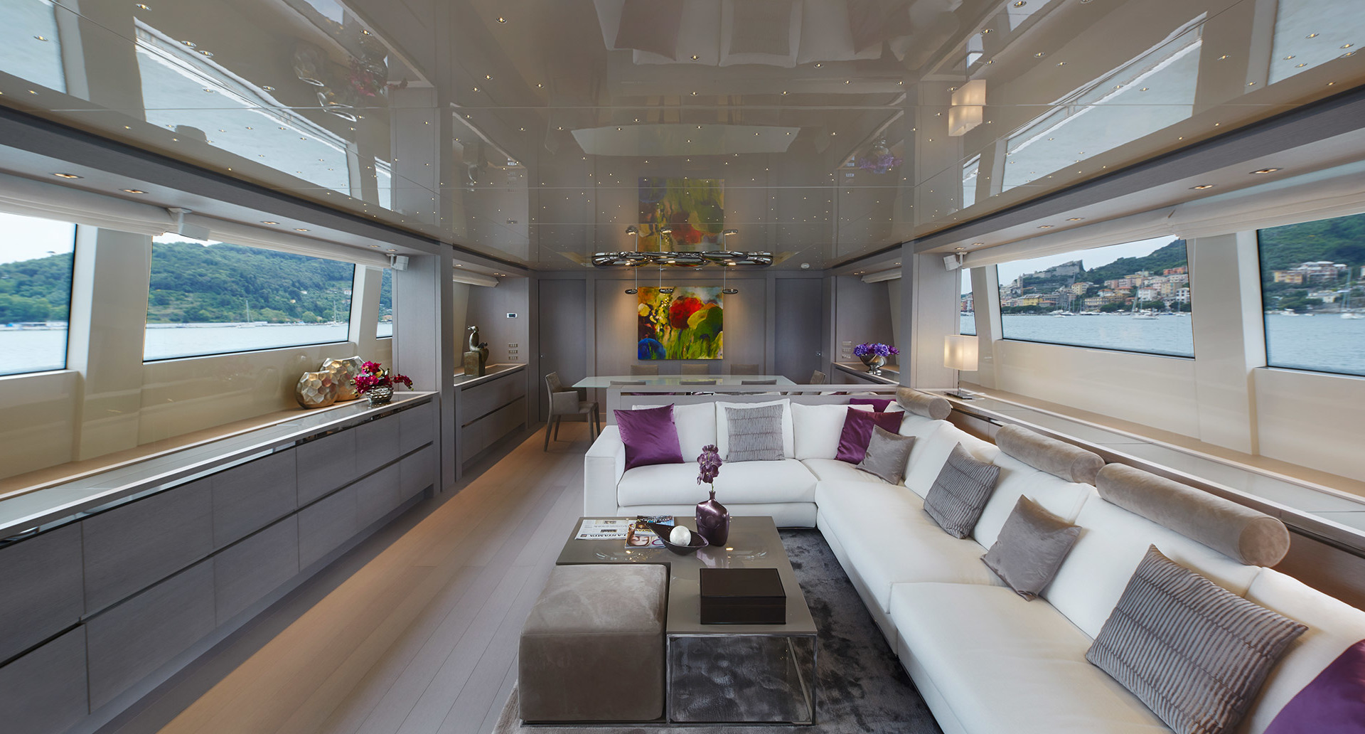 Luxury yacht salon with white sectional sofa, purple accents, panoramic windows, and elegant pendant lighting over a coffee table.