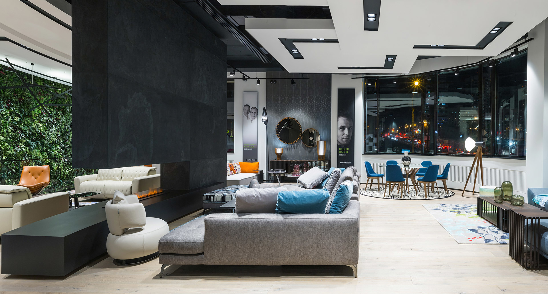 Modern showroom with recessed ceiling lights, floor lamp on tripod, gray sofa, fireplace, green living wall, and city view at night