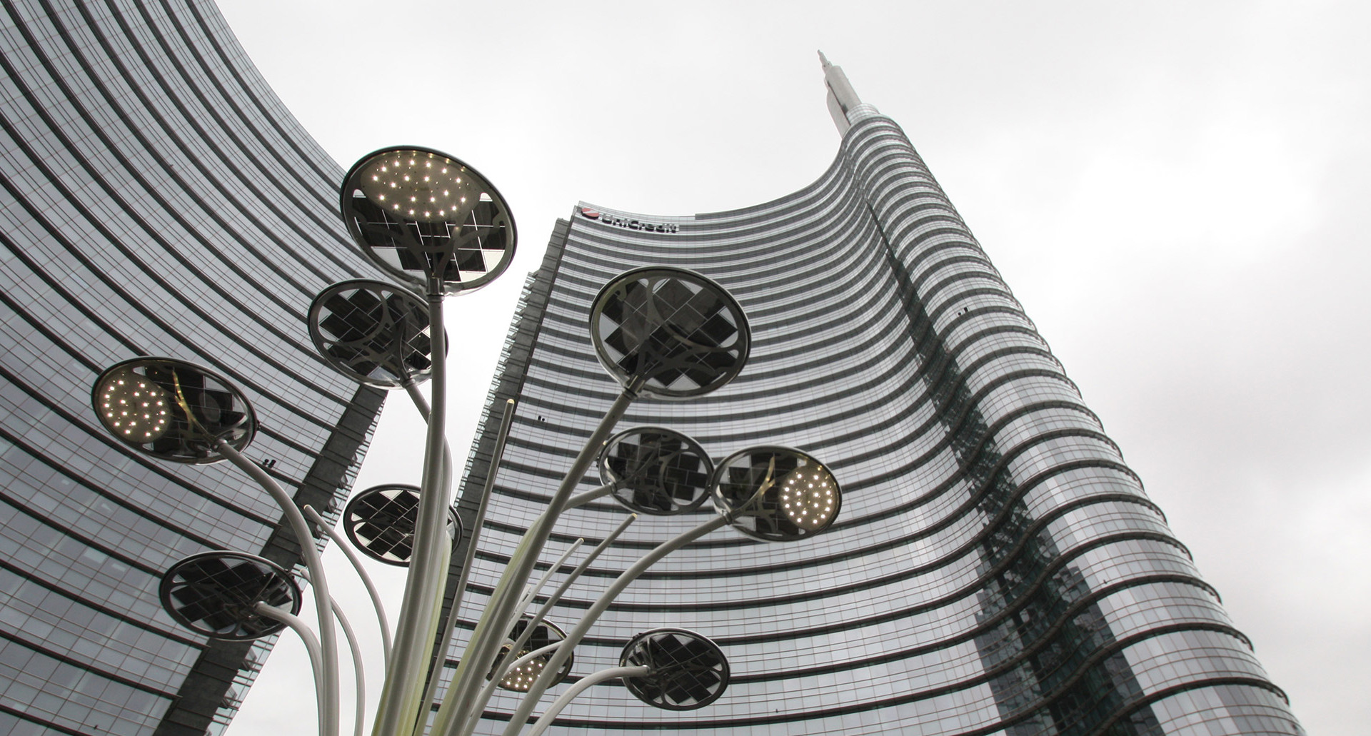 Artemide outdoor street lamps with circular disc heads on tall poles in front of the curved glass UniCredit Tower, Milan.