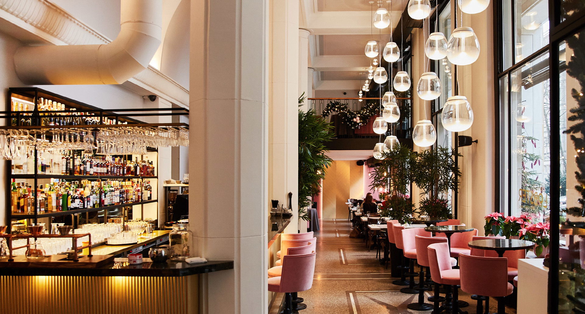 Elegant restaurant interior with cascading glass pendant lights over pink velvet chairs, warm bar area with hanging glassware in background