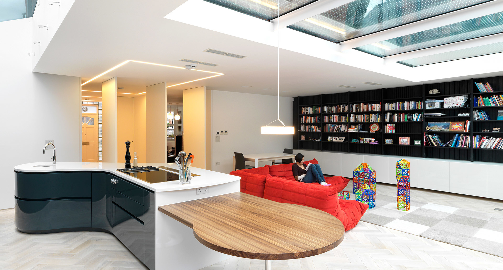 Modern open-plan living space with Artemide pendant light over kitchen island, skylight ceiling, red sofa, and floor-to-ceiling bookshelf.