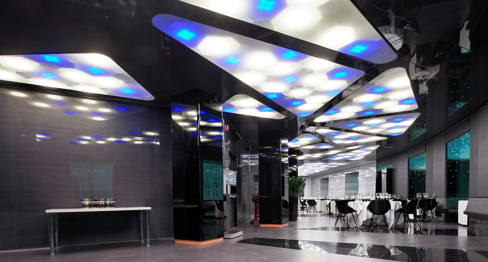 Modern lobby with geometric ceiling panels featuring white and blue LED lighting, reflecting on polished dark floors, with dining area in background