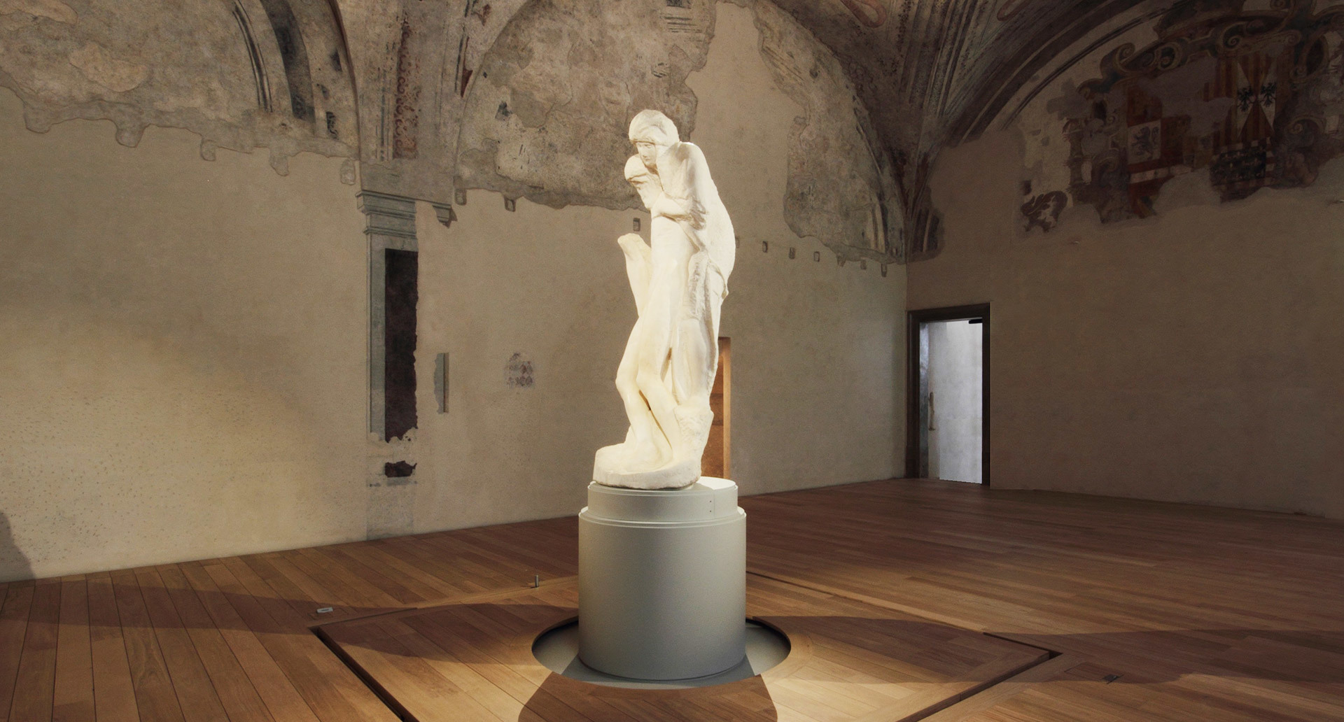 Classical marble statue on a cylindrical pedestal with floor lighting in a historic vaulted hall with frescoed walls.