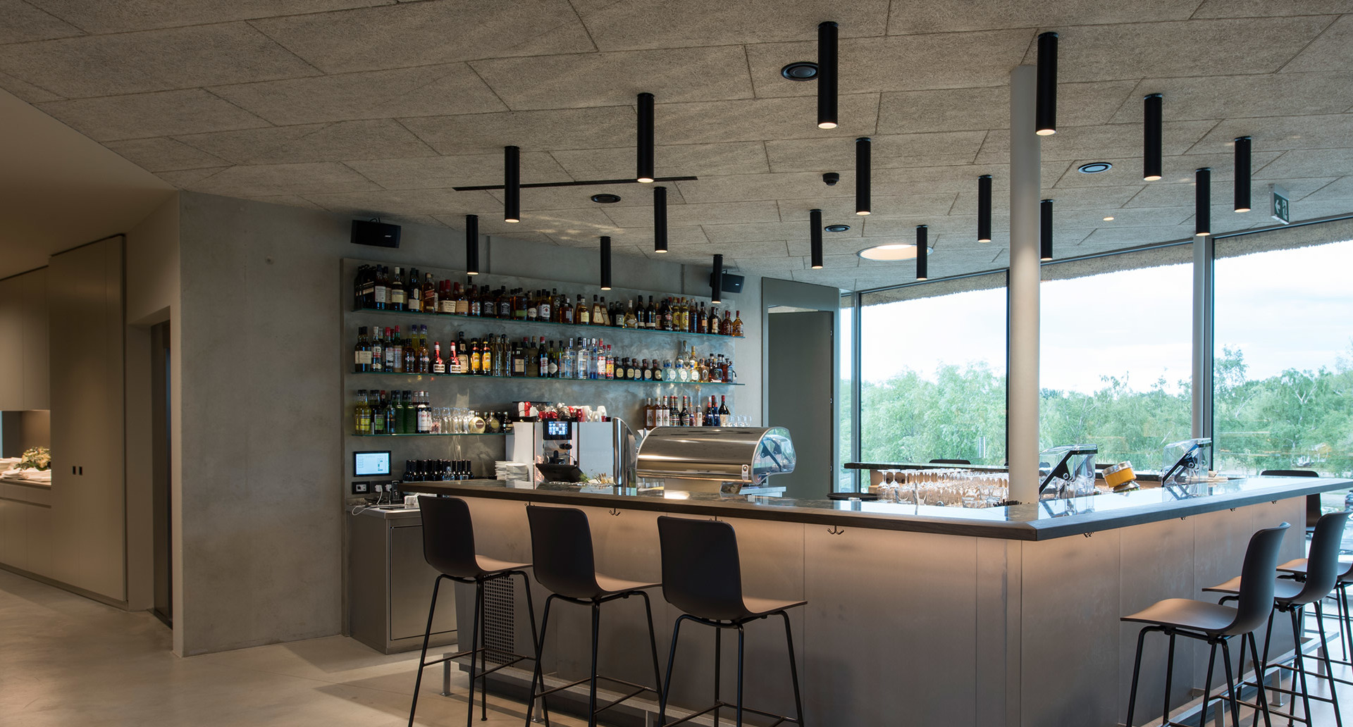 Modern bar interior with black cylindrical pendant lights arranged on ceiling, illuminating a sleek counter with bar stools and bottle display.