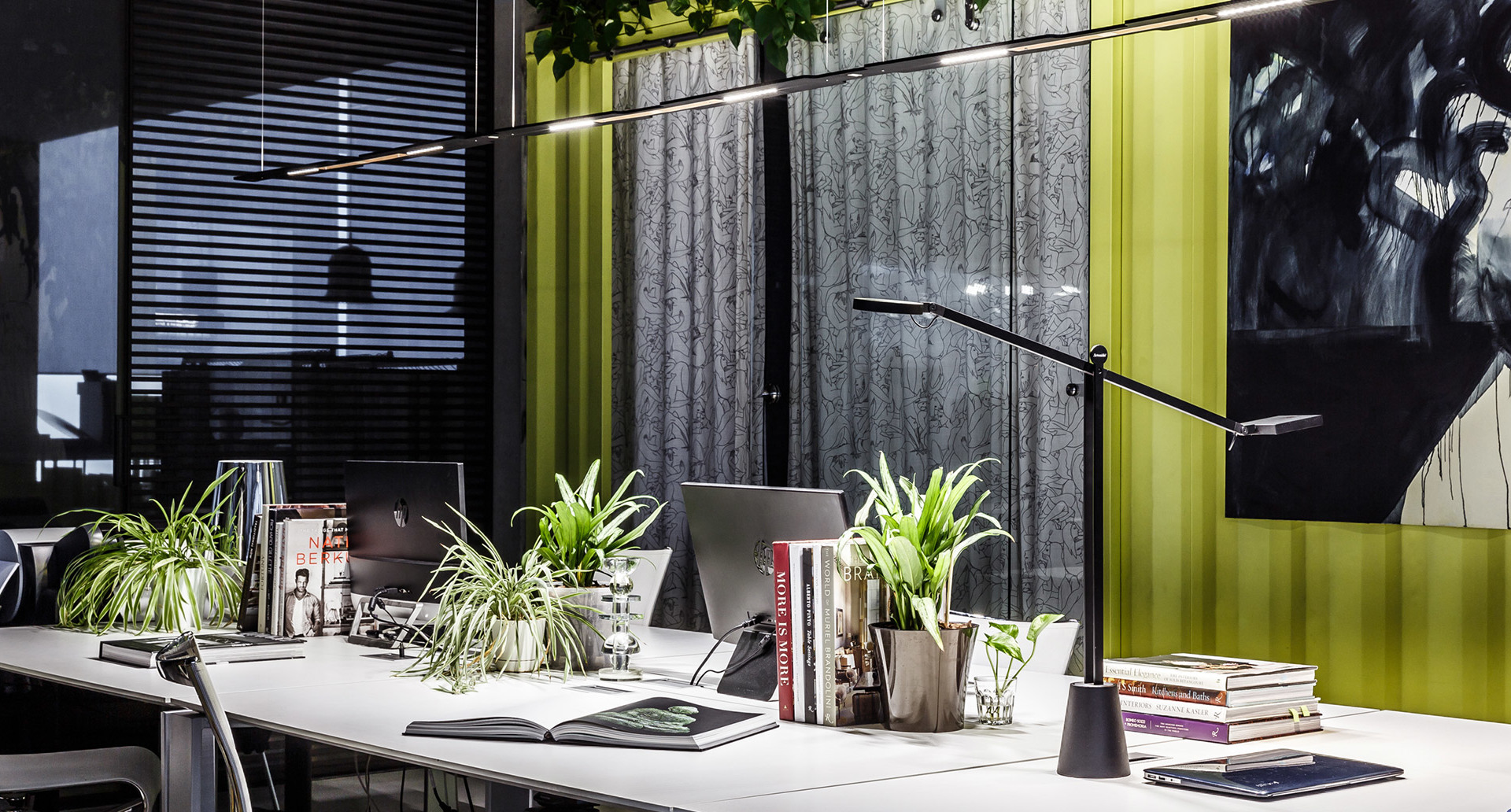 Modern office desk with black adjustable task lamp and suspended linear lamp, surrounded by green plants and books.