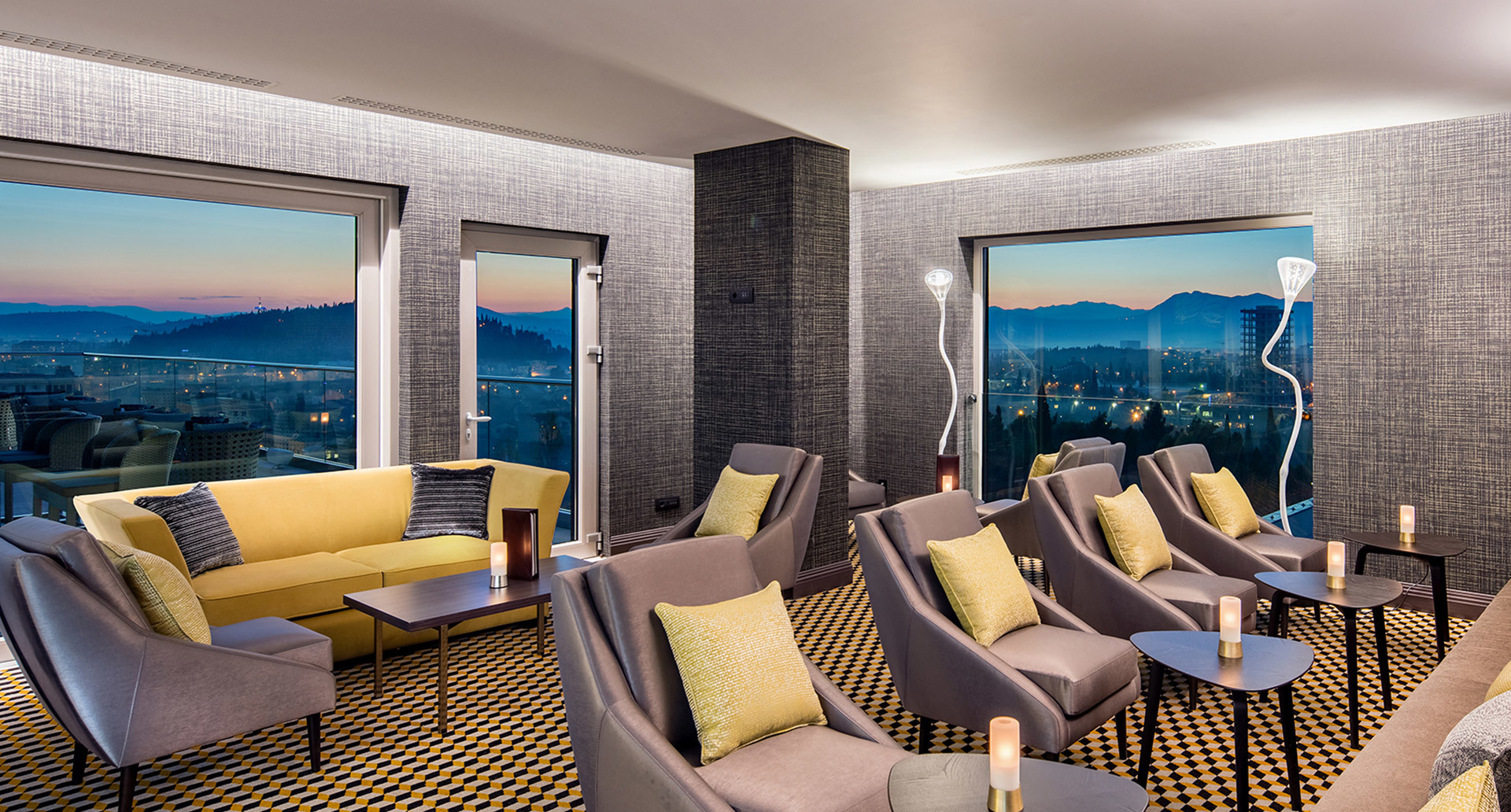 Elegant hotel lounge at dusk with Artemide floor lamps, yellow sofas, grey armchairs, patterned carpet, and panoramic city views