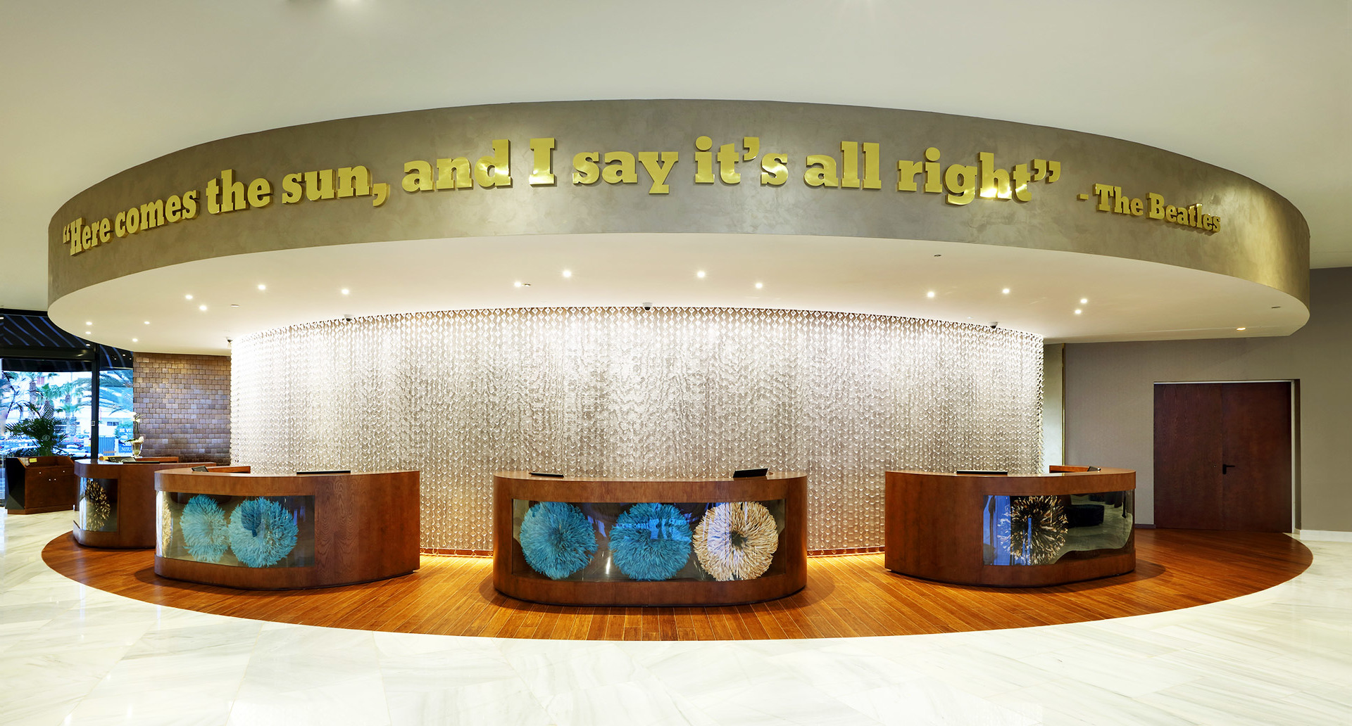 Hotel lobby with curved reception desk, decorative wall, and illuminated ring ceiling feature inscribed with The Beatles quote.
