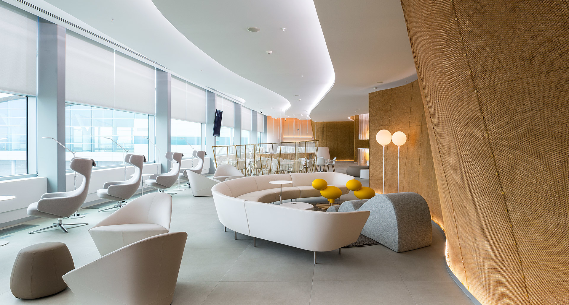 Modern lounge with curved white sofas, yellow accents, Artemide floor lamps, and elegant indirect ceiling lighting creating a warm ambiance.