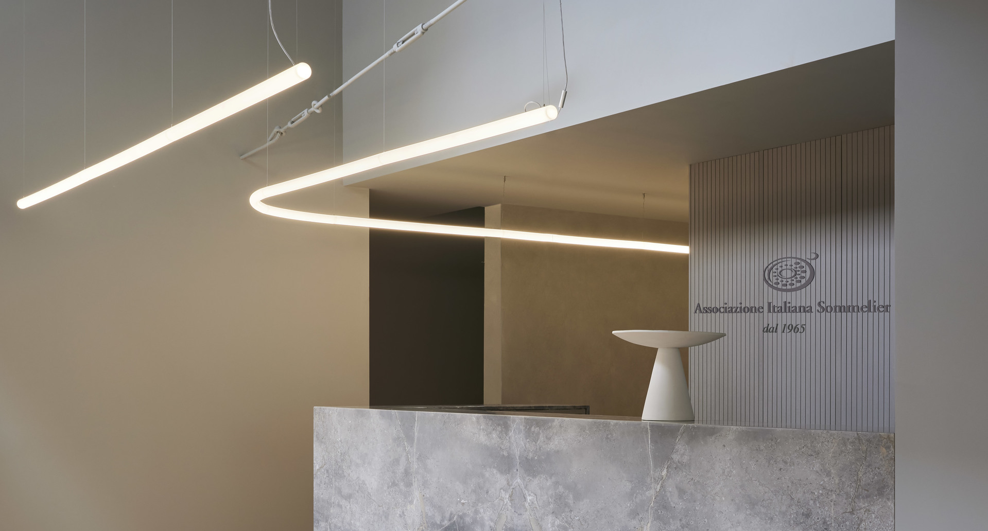 Artemide curved LED pendant light forming an open loop shape, suspended over a marble reception desk in a minimalist interior.