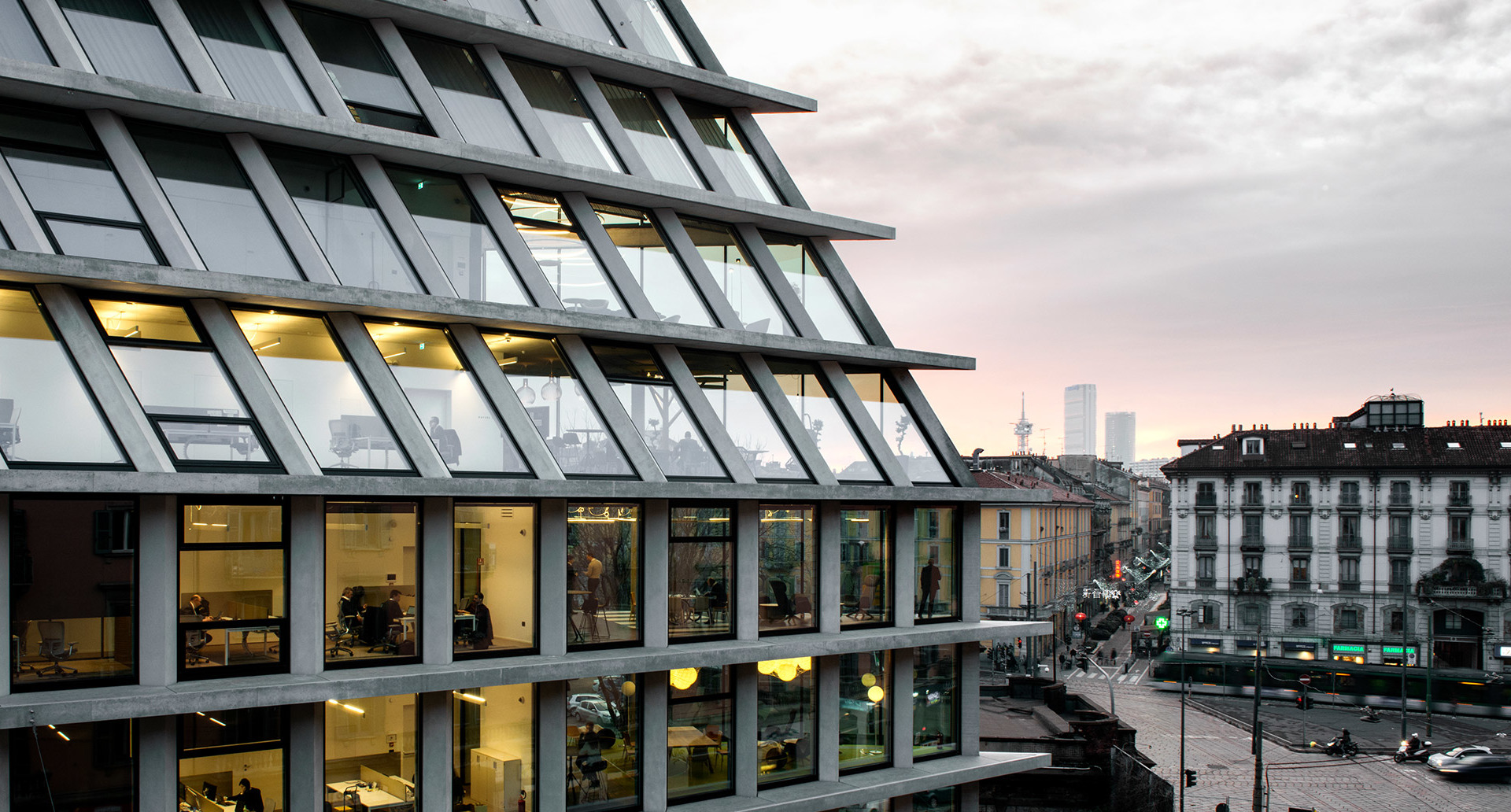 Modern office building facade with diamond-shaped glass panels illuminated at dusk, overlooking a Milan city street intersection.