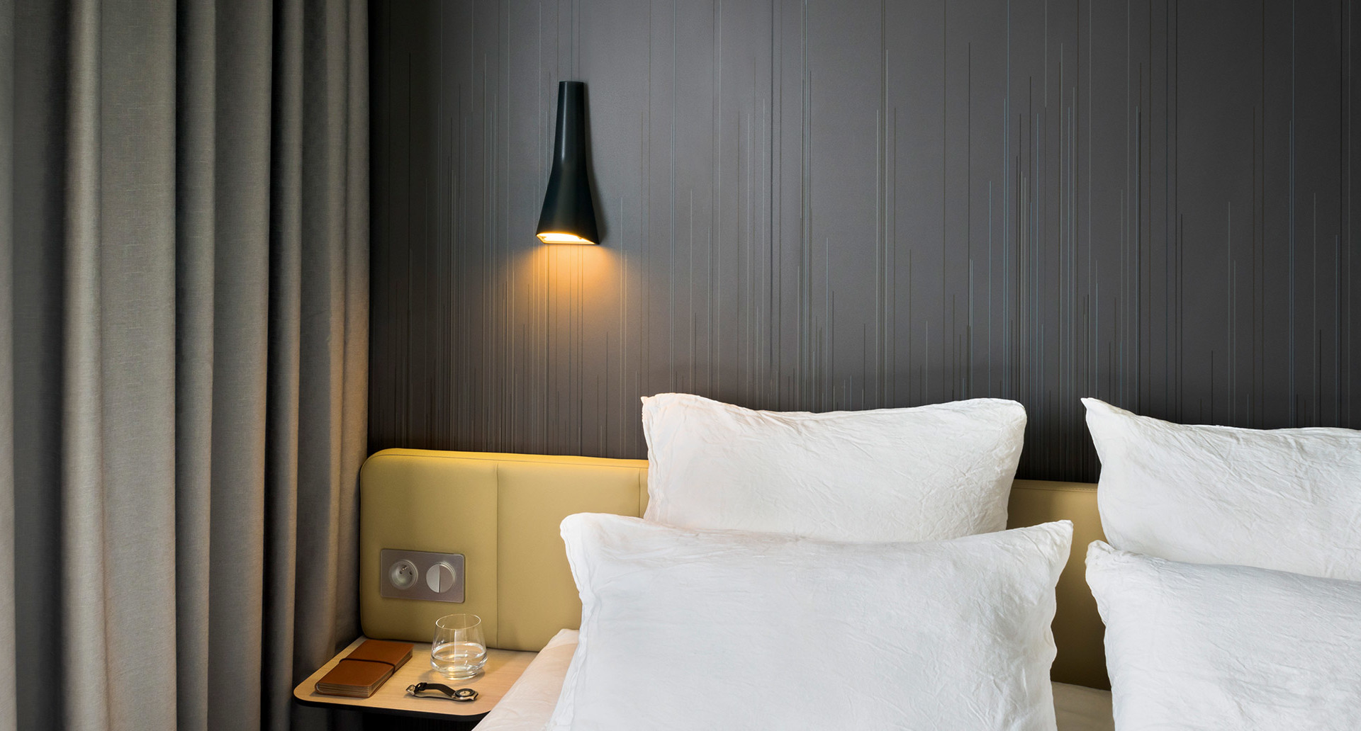 Black conical wall lamp casting warm downward light above a hotel bed headboard with white pillows and grey wood panel.