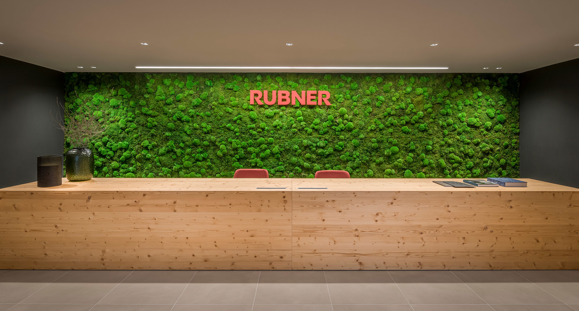 Rubner reception desk in natural wood with green moss wall backdrop, illuminated by recessed ceiling spotlights.