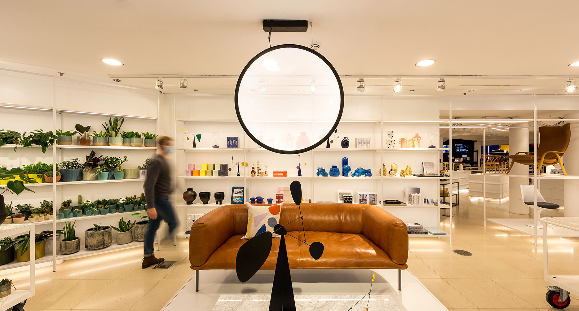 Artemide circular pendant lamp illuminates a modern showroom with leather sofa, colorful shelves, plants, and a mobile sculpture.