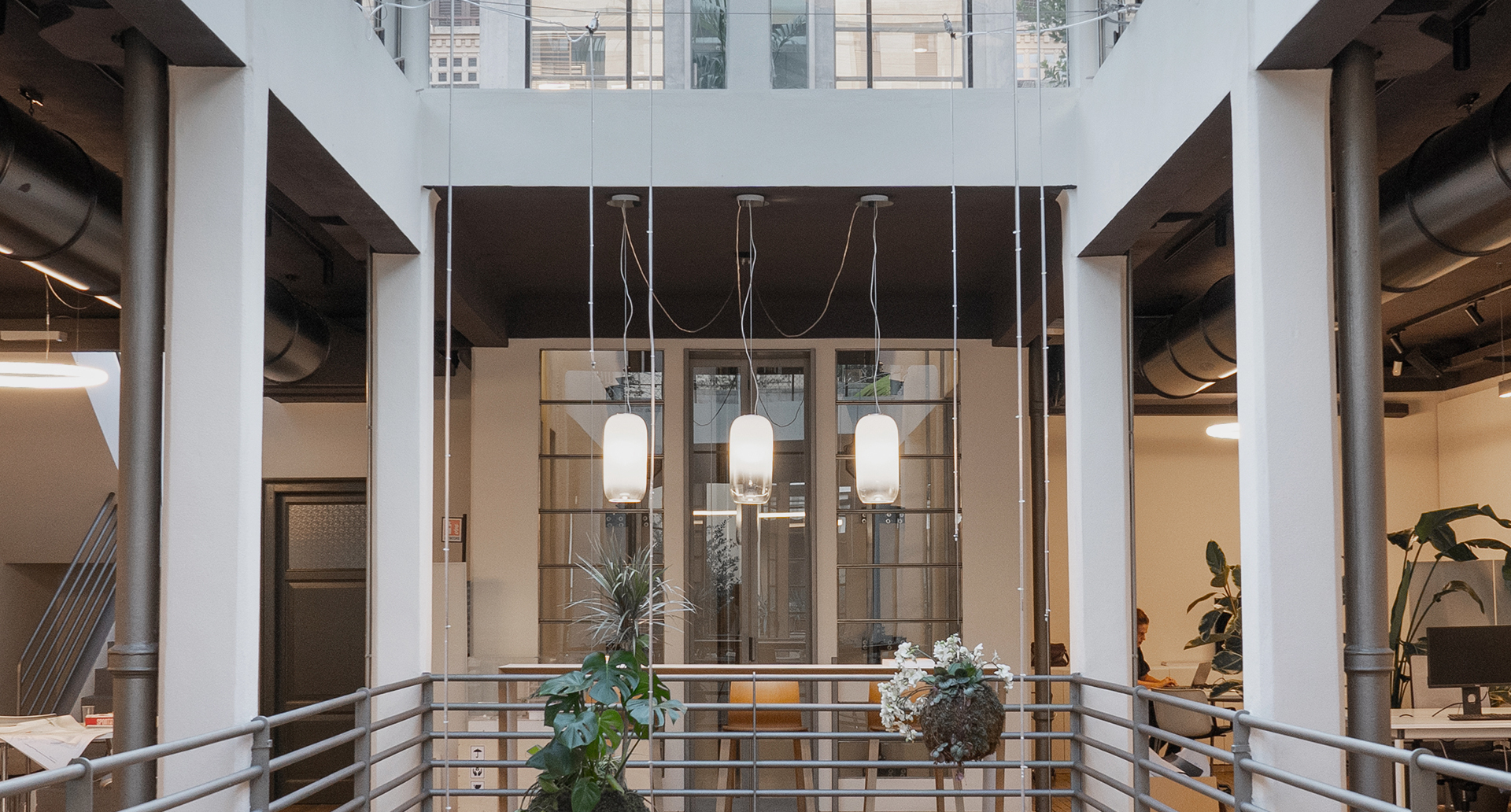 Three white cylindrical pendant lamps hanging from high ceiling in a modern open-plan office atrium with columns and glass walls