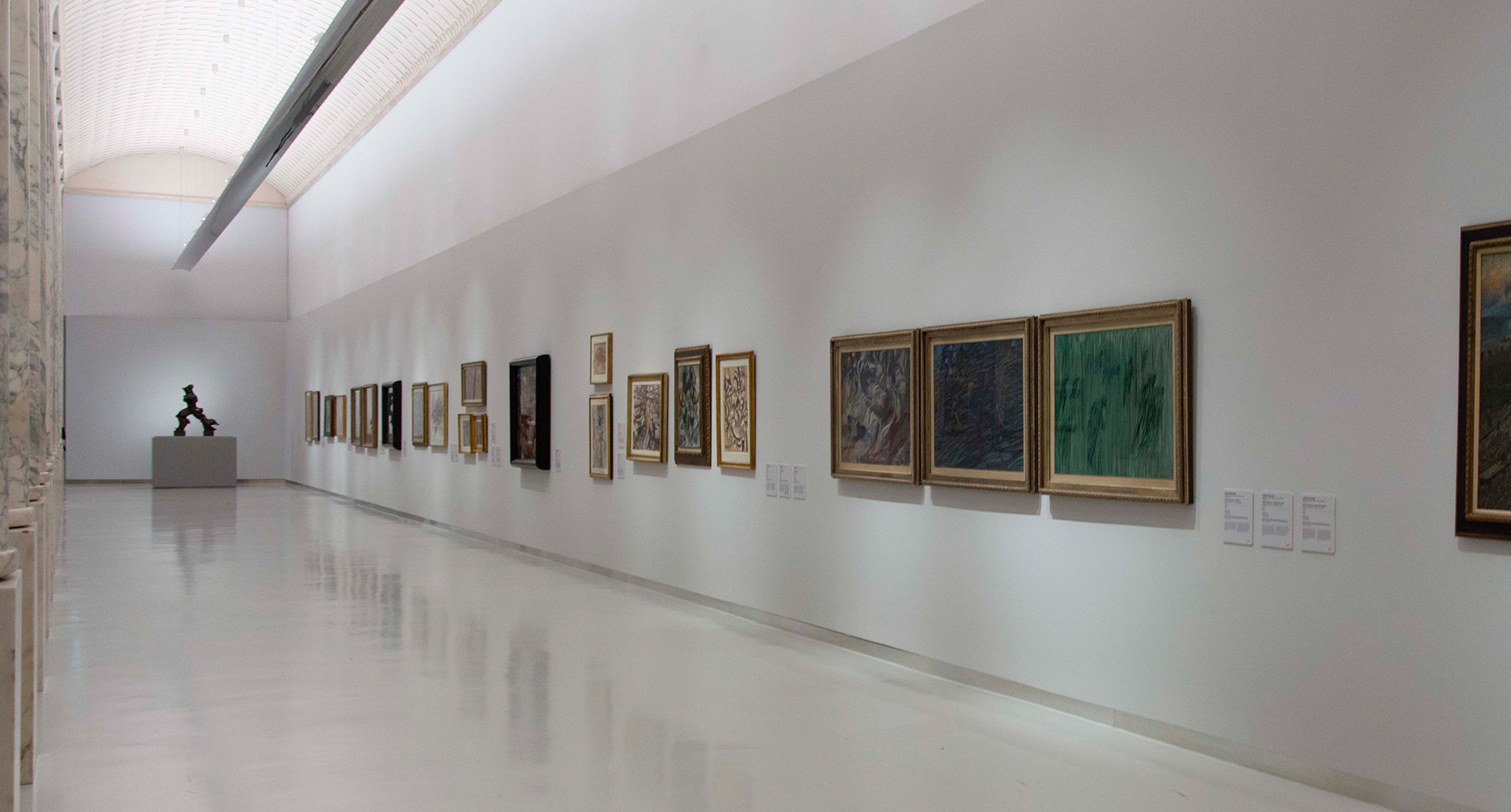 Art gallery corridor with recessed ceiling lighting illuminating paintings on white walls and a sculpture at the end.