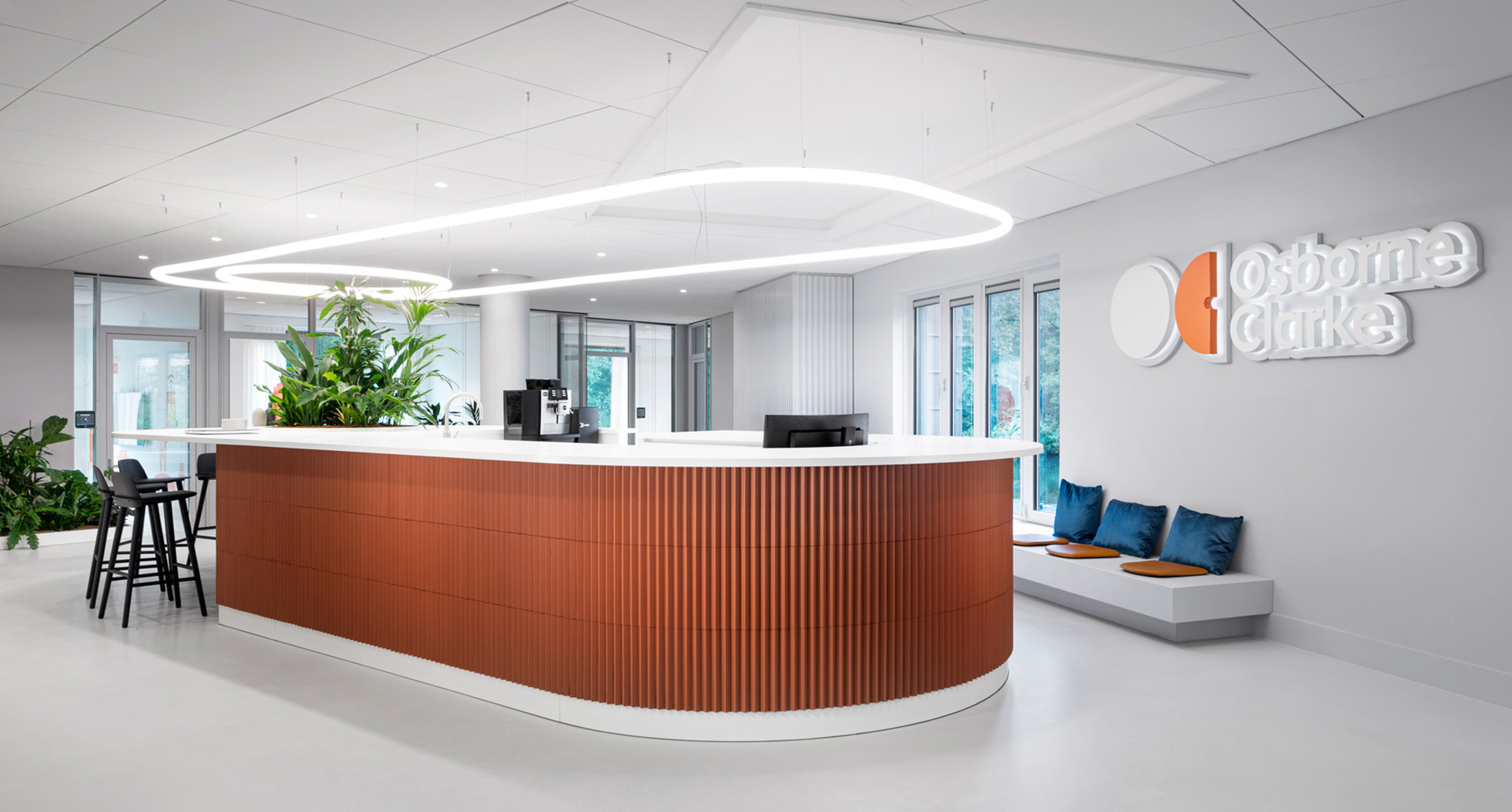 Osborne Clarke office reception with Artemide oval ring pendant light illuminating a curved wood-paneled desk and green plants.