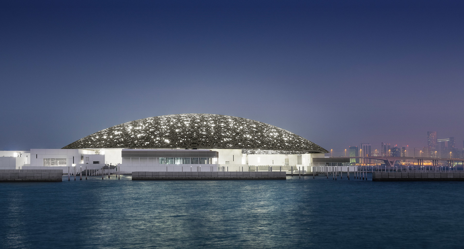Louvre Abu Dhabi museum at dusk, its iconic illuminated dome reflecting on calm waters with city skyline in background