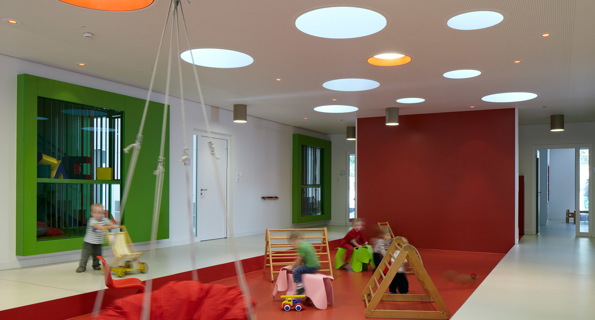 Colorful children's playroom with large circular ceiling lights illuminating kids playing on red floor with toys and swings.