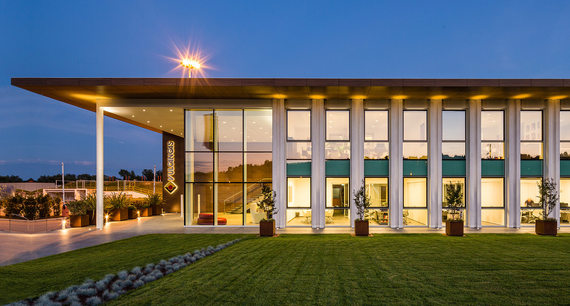 Modern glass-facade building illuminated at dusk, featuring warm interior lighting and recessed ceiling fixtures on an extended flat roof.
