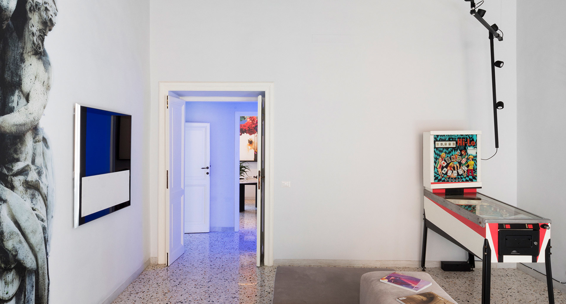 Modern white room with pinball machine, wall art, TV, and a black floor lamp with multiple adjustable spotlights on a vertical pole
