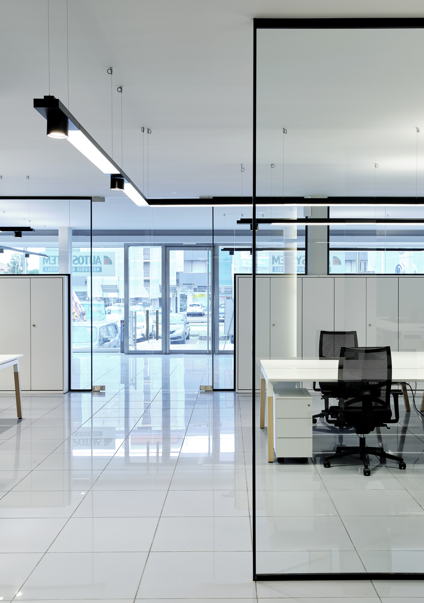 Modern office with glass partitions and black pendant linear LED lights illuminating white workstations and polished tile floors.