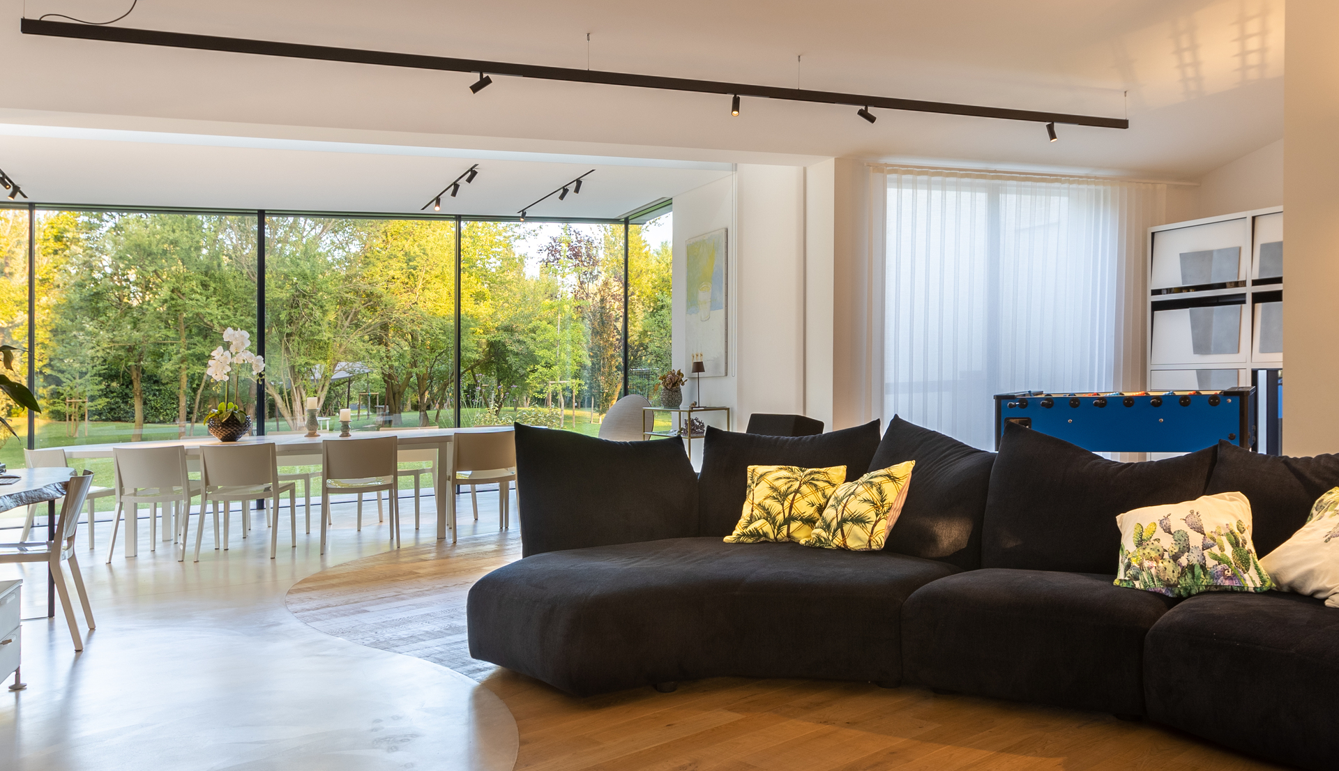 Modern open-plan living room with track lighting on ceiling, large black curved sofa, floor-to-ceiling windows overlooking a lush garden