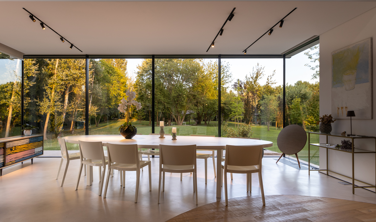 Modern dining room with oval white table, beige chairs, and black track lighting on ceiling, overlooking a lush garden through floor-to-ceiling windows.