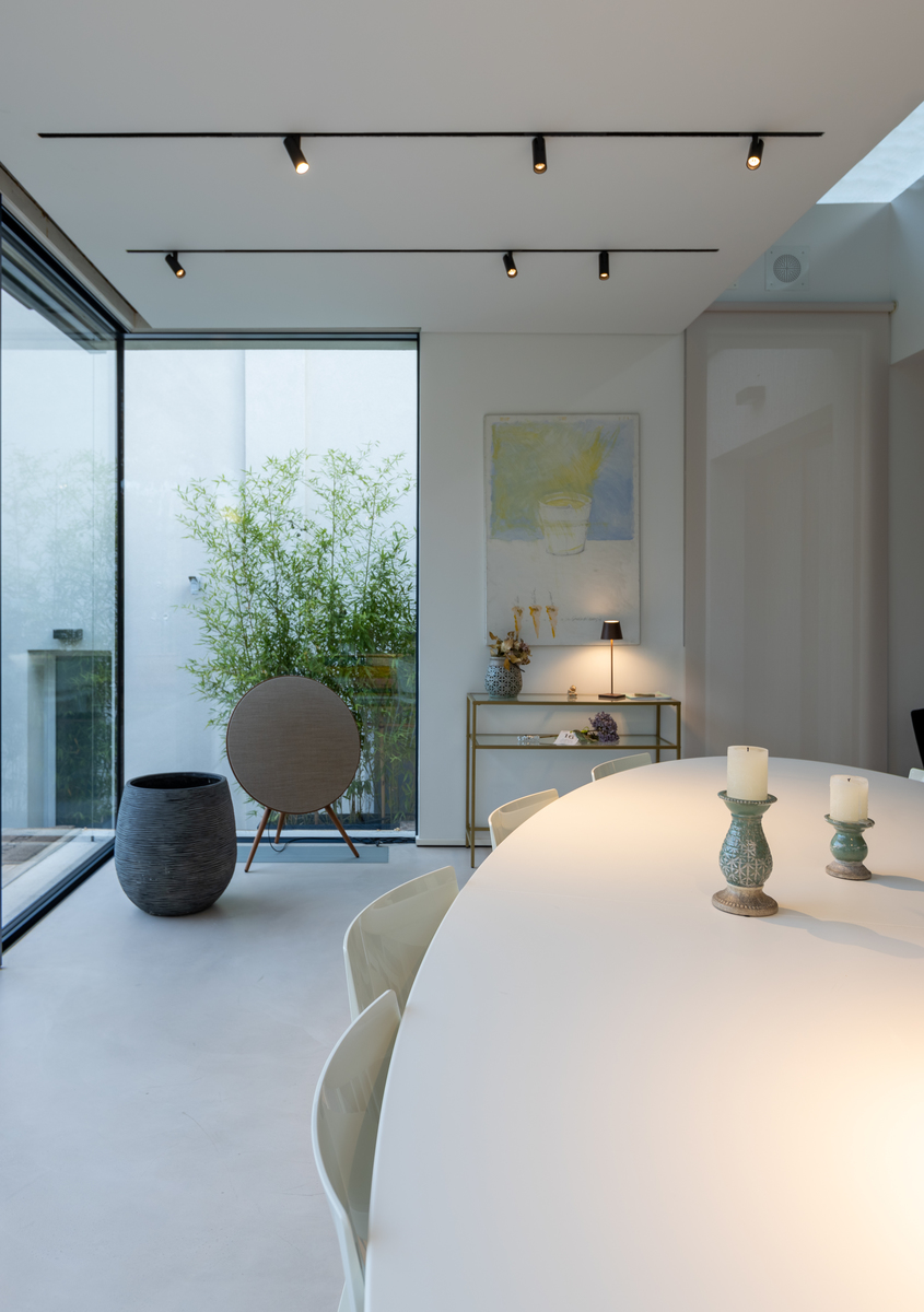 Modern dining room with white oval table, track lighting on ceiling, floor-to-ceiling glass wall opening to a courtyard garden