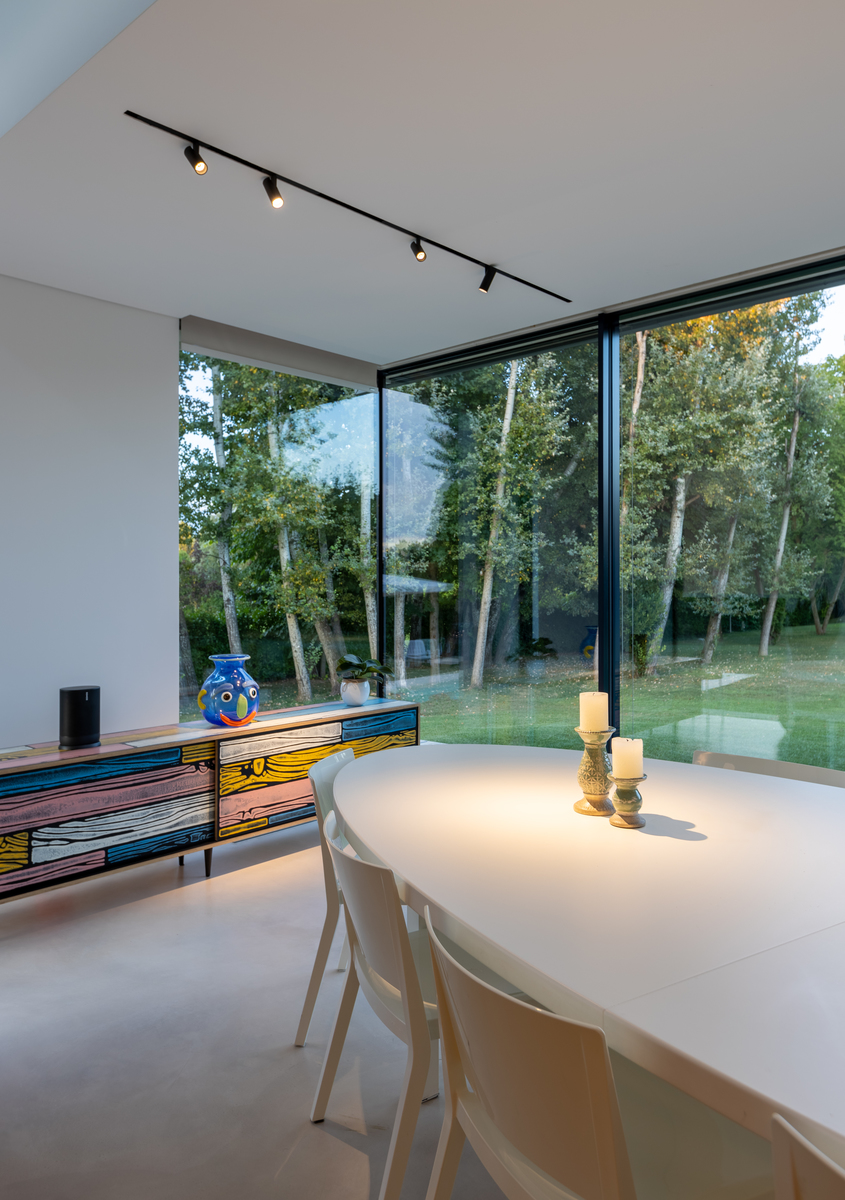 Dining room with white round table, Artemide track spotlights on ceiling, floor-to-ceiling windows overlooking a garden