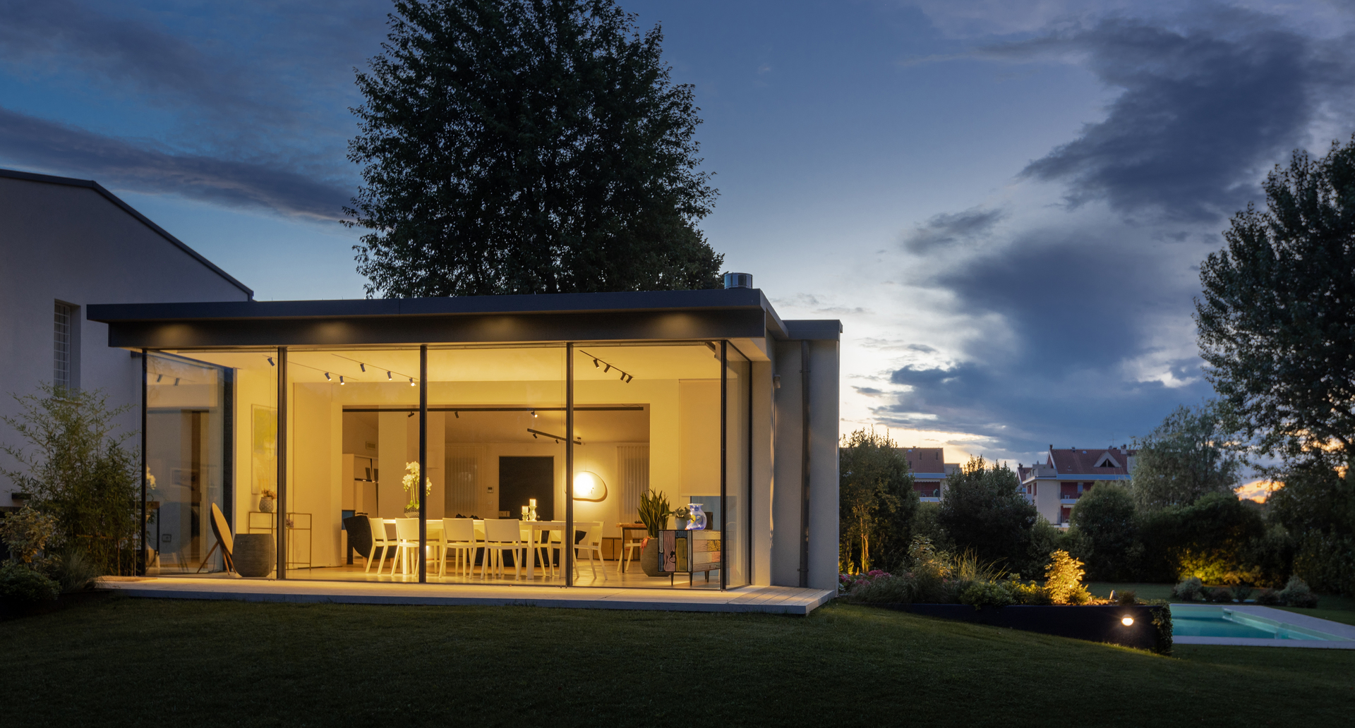 Modern glass-walled house at dusk, warmly lit interior with track spotlights and a pendant lamp visible through floor-to-ceiling windows