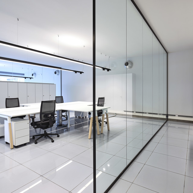 Modern office workspace with white desks, mesh chairs, and track lighting on ceiling, separated by a floor-to-ceiling glass partition.