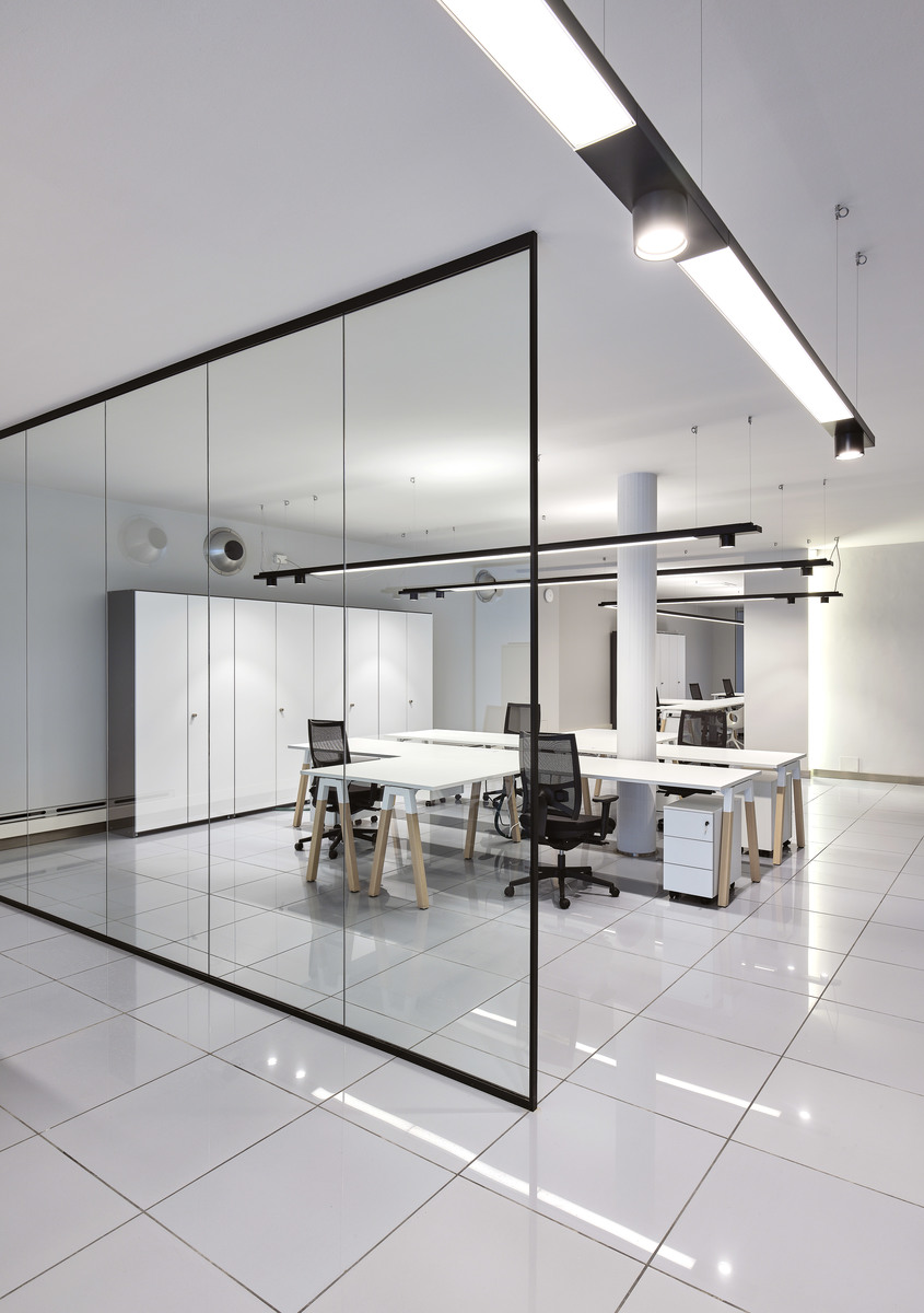 Modern open office with glass partition walls, white workstations, and Artemide linear LED ceiling lights providing uniform illumination