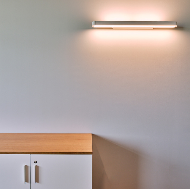 White rectangular wall-mounted LED light fixture casting warm upward glow on wall above a wooden storage cabinet.