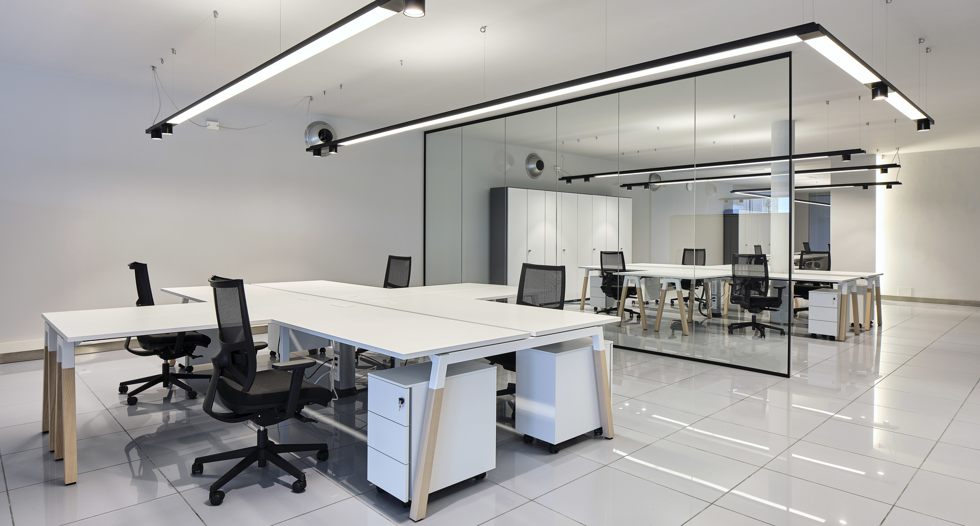 Modern open-plan office with white workstations, mesh chairs, and Artemide linear LED ceiling lights illuminating glass-partitioned meeting room.
