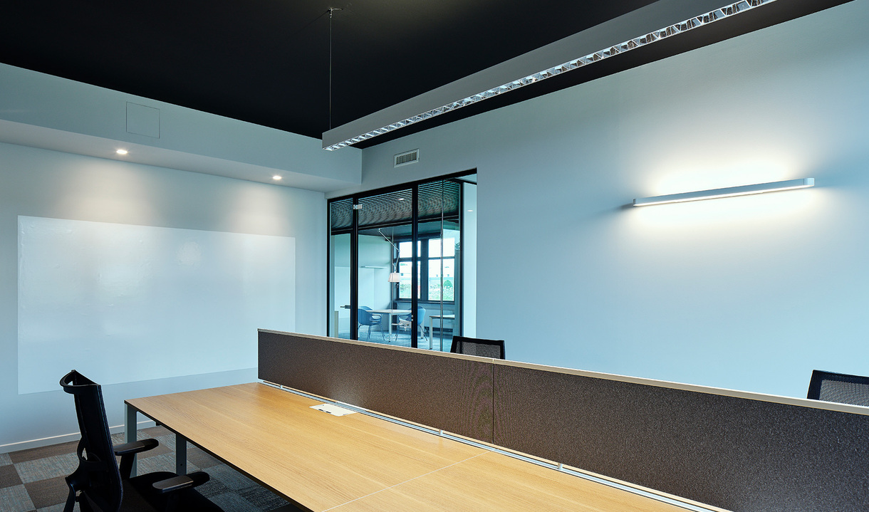 Modern office workspace with workstations, dark ceiling, pendant linear light, recessed spotlights, and a curved wall-mounted lamp on blue-grey wall.