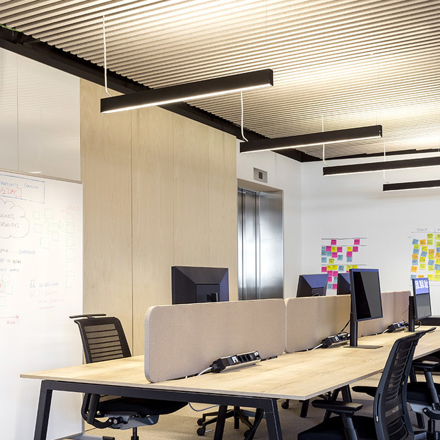 Modern office with black linear pendant lights illuminating rows of workstations with monitors, dividers, and a whiteboard wall.