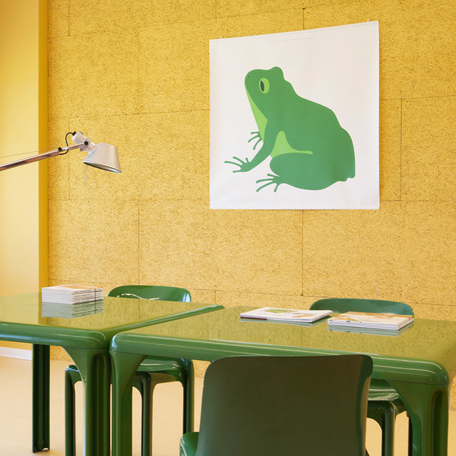 Artemide Tolomeo table lamp on a green desk with matching chairs, yellow cork wall, and a green frog artwork in the background
