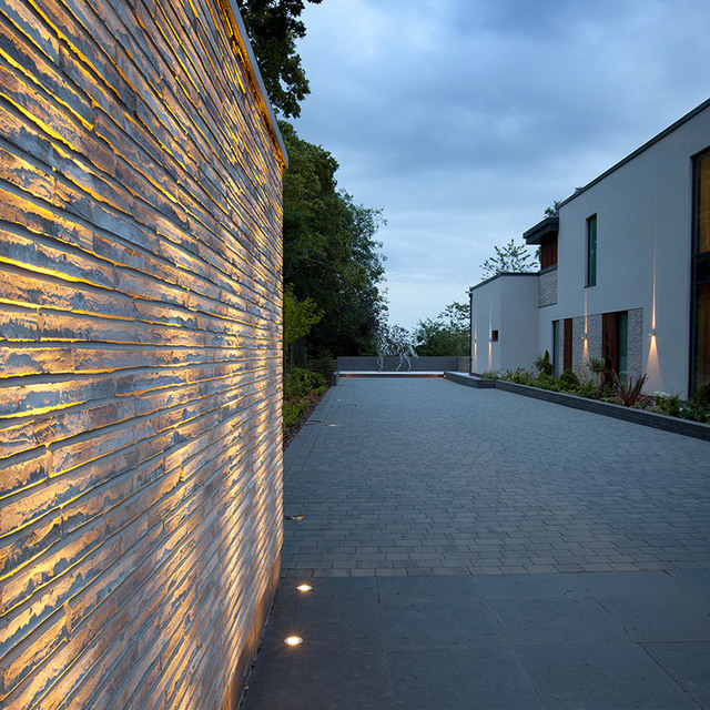 Driveway at dusk with warm LED lights grazing a textured stone wall and recessed ground spotlights illuminating the path to a modern house