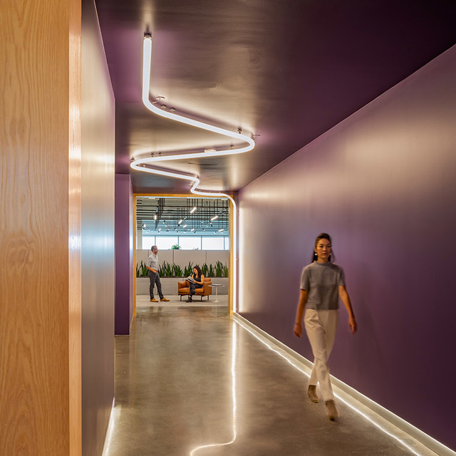 Modern office corridor with purple walls featuring a zigzag LED tube ceiling light and floor-level strip lighting, with people walking.