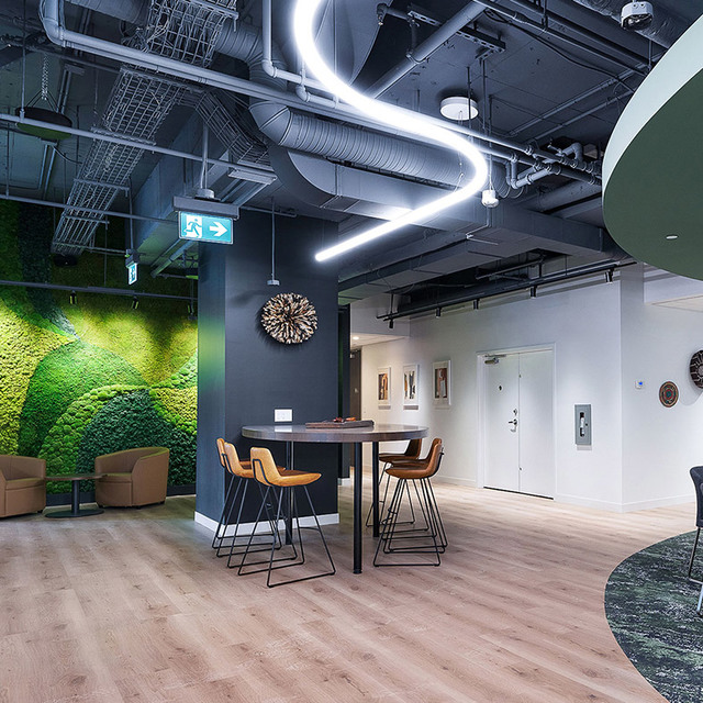 Modern office lounge with a sinuous LED linear pendant light, moss green wall, exposed ducts, high bar table, and warm wood flooring