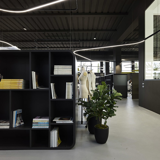 Modern office interior with Artemide curved LED pendant lights on dark ceiling, black bookshelf, mannequins, and potted plants.