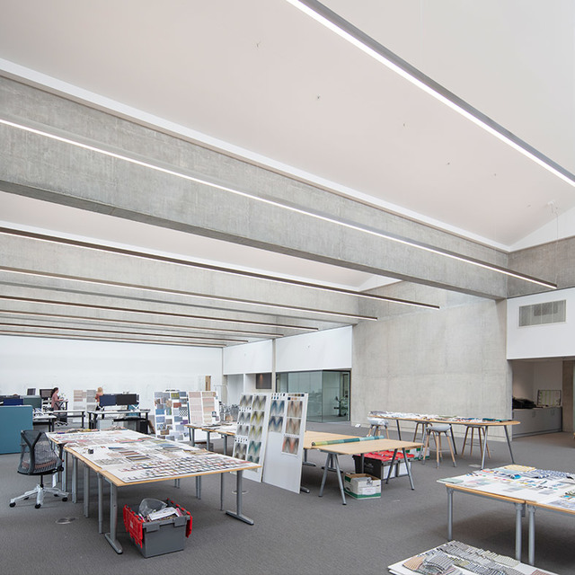 Open-plan studio workspace with linear LED ceiling lights illuminating work tables covered with design drawings and material samples.