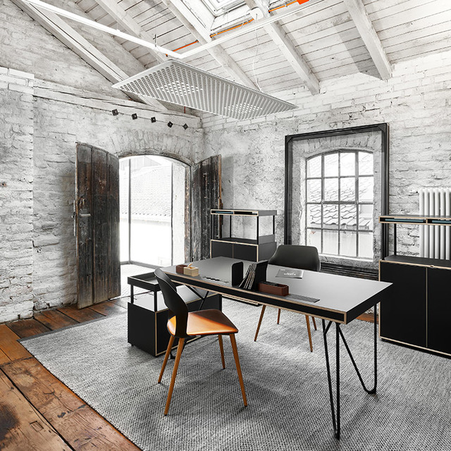 Industrial-style office with exposed brick walls, skylight, track lighting, black desk with hairpin legs, and modern chairs on a grey rug