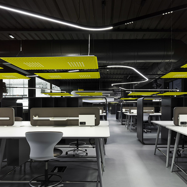 Modern open-plan office with curved white LED linear pendant lights, yellow acoustic ceiling panels, and white workstations on dark flooring