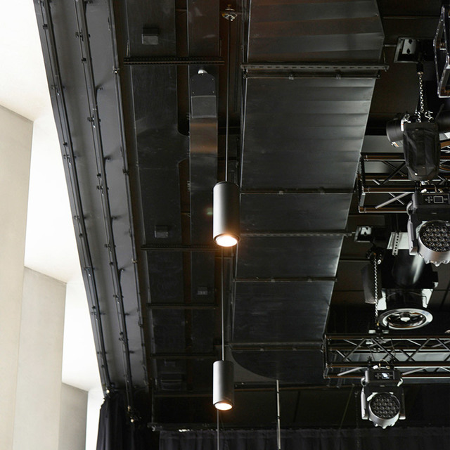 Two cylindrical black pendant lights hanging from a dark industrial ceiling with metal trusses and stage lighting equipment.